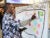 A black man with dredlocks, a blue patterned shirt, and a white undershirt, places a pink sticky note on a white board that asks “What could the U.S. look like post-reparations?”. The whiteboard is decorated with stars, a light bulb, a green flag, dotted lines, and other post-its.
