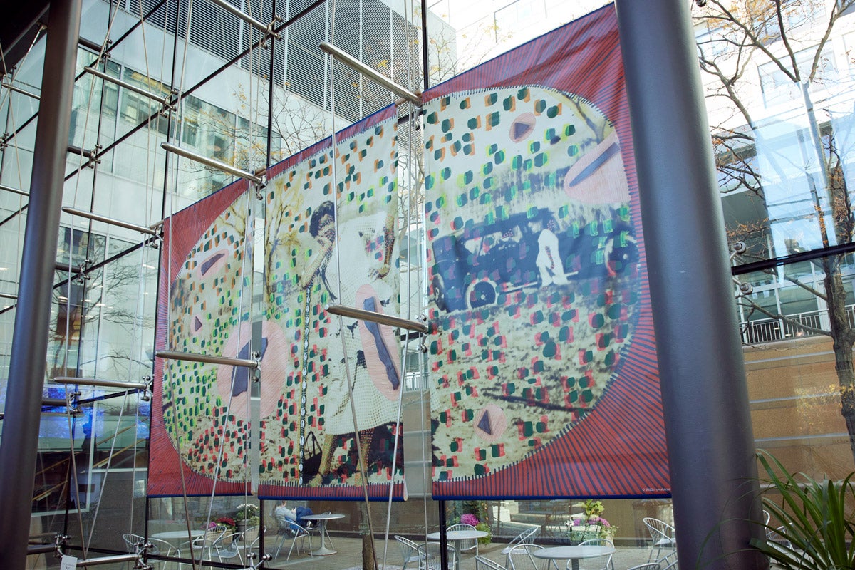 A three-paneled artwork hangs in an atrium. The artwork is a collage of photographs: a black woman leaning on an implied fence, an old car, and other indistinguishable objects. The artwork is in an oval and has a red and black sunburst border.