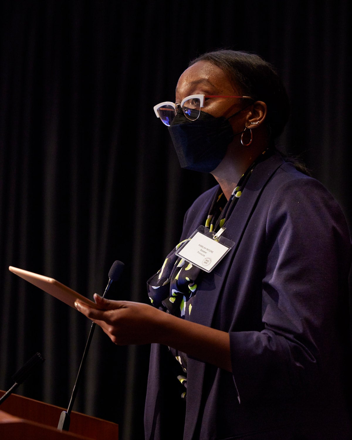Kamilah Moore wears a black facemaks, bright white cat-eyed glasses and holds an ipad while speaking at a podium