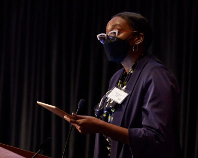 Kamilah Moore wears a black facemaks, bright white cat-eyed glasses and holds an ipad while speaking at a podium