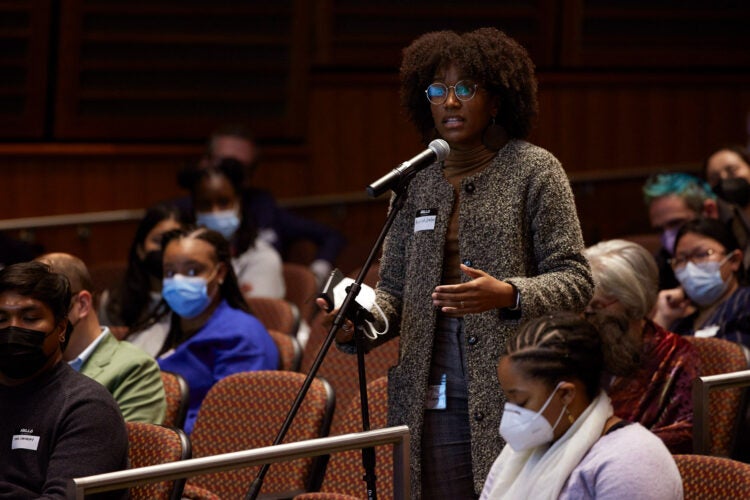 A black female wearing a long sweater cardigan, glasses and natural curly hair, asks a question at a microphone in an auditorium.