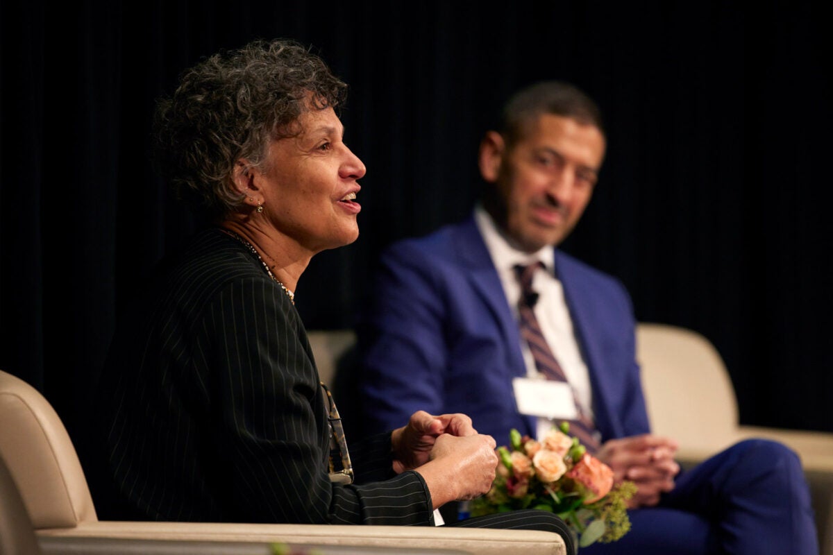 Mary T. Bassett sits in a cream armchair and speaks to an audience. Sandro Galea sits on her left in his own chair in the background.