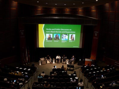 Four panelists sit in armchairs at the front of an auditorium. Behind them is a large screen with yellow and green color blocking, the title of the panel, and headshots of the four panelists (unlegible.)