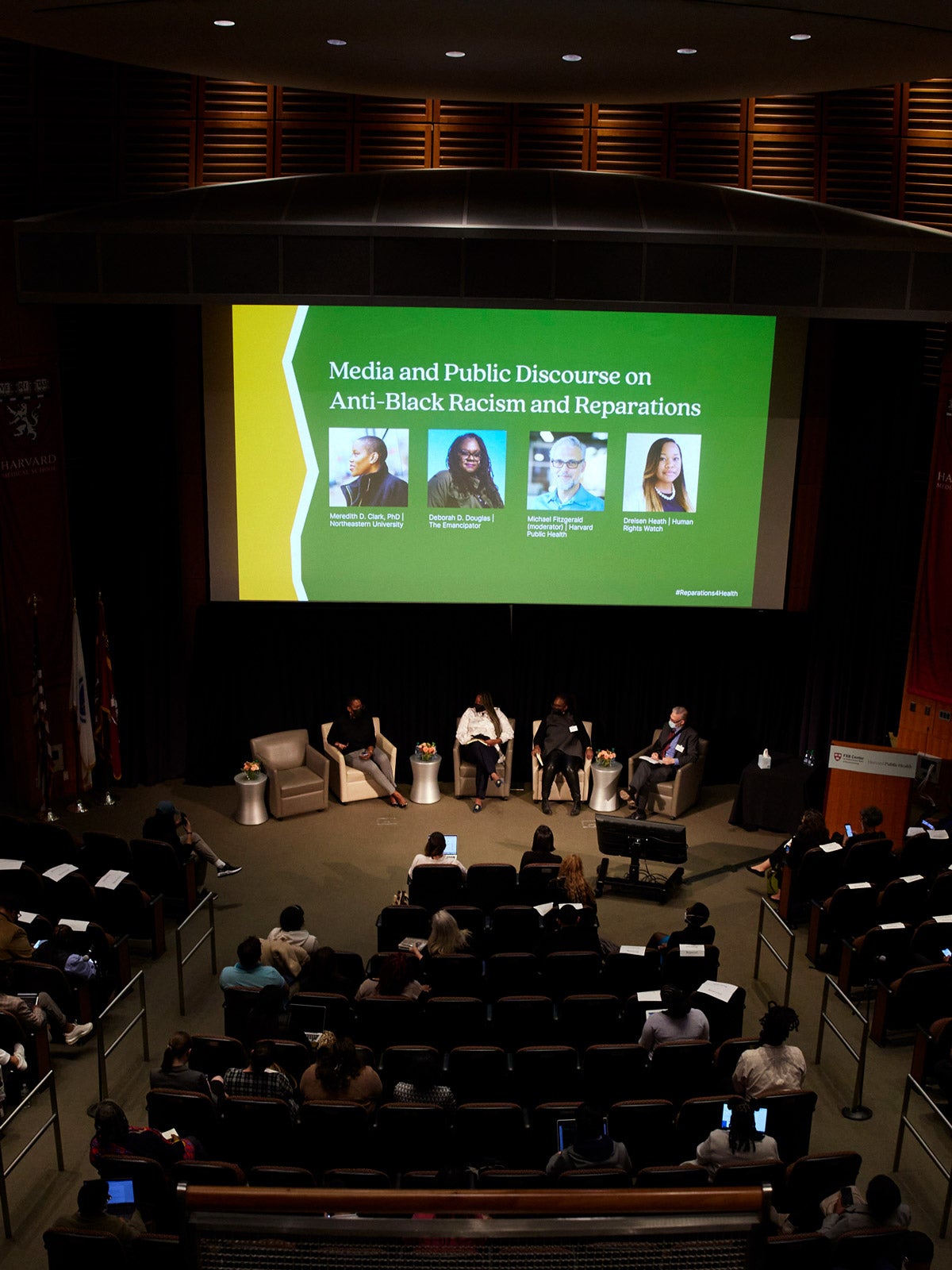 Four panelists sit in armchairs at the front of an auditorium. Behind them is a large screen with yellow and green color blocking, the title of the panel, and headshots of the four panelists (unlegible.)