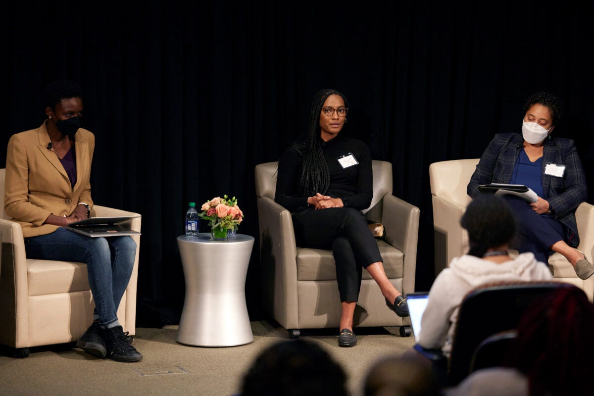 Three black women sit in cream armchairs on a stage addressing an audience. . The middle woman is speaking.