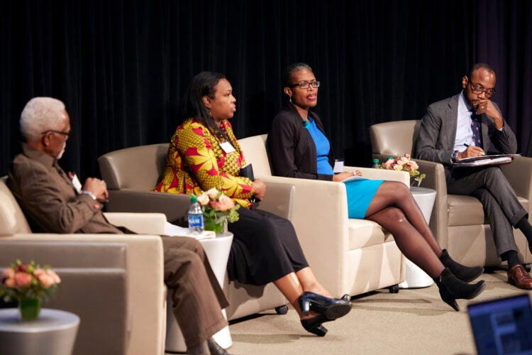 Two black men and two black women sit in cream armchairs on a stage addressing an audience. The third person (black woman) is speaking.