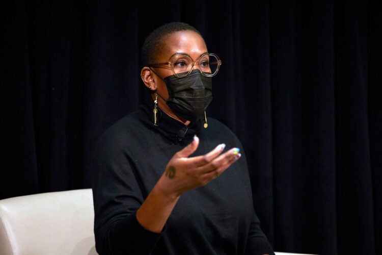 A Black female sits on stage speaking with her right hand slightly raised. She wears a black turtleneck, glasses, and a black facemask.
