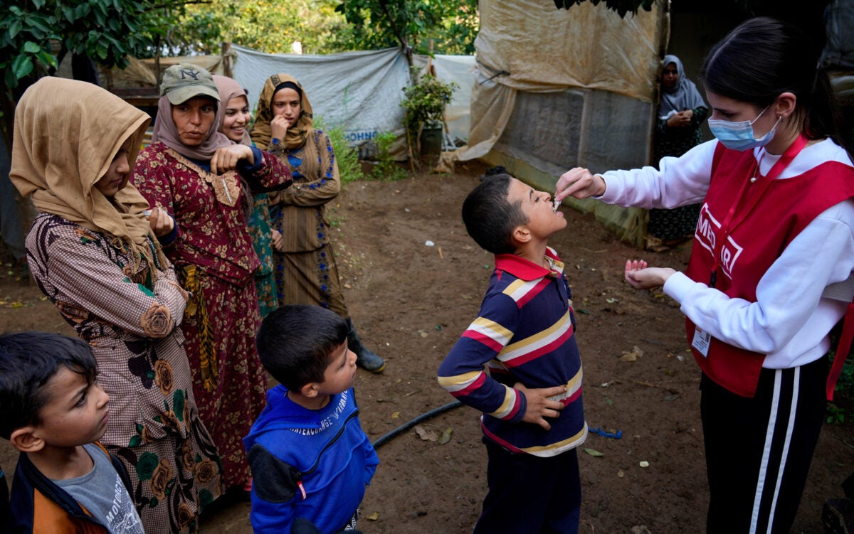 A Syrian child wearing a striped shirt receives an oral Cholera vaccine from a female aid worker in North Lebanon. Two other young boys look on as well as a group of four Syrian women.