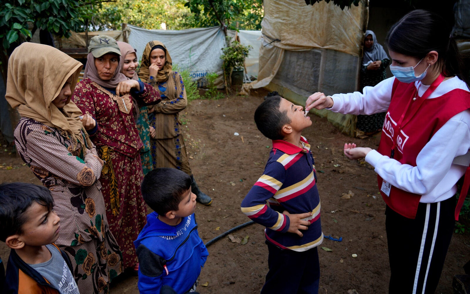 A Syrian child wearing a striped shirt receives an oral Cholera vaccine from a female aid worker in North Lebanon. Two other young boys look on as well as a group of four Syrian women.