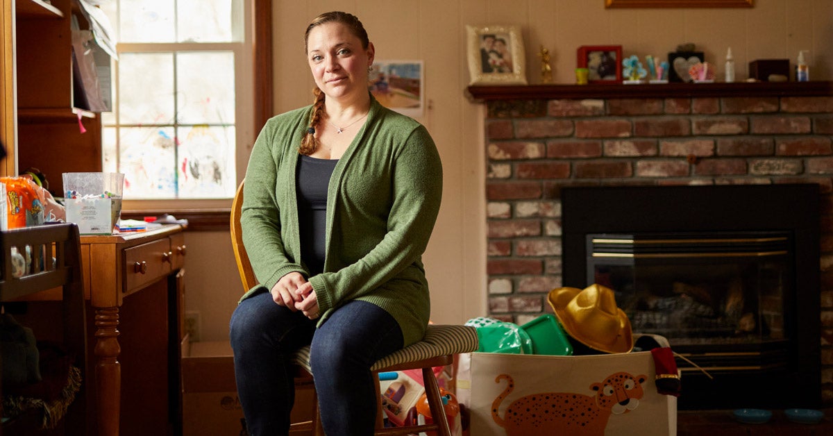 A white middle-aged woman sits in a living room. She wears a green cardigan, tank top and jeans, and her hair is braided in a single braid; hands clapsed in her lap. Surrounding her are children's toys, a bin on the floor and a fireplace with photos.