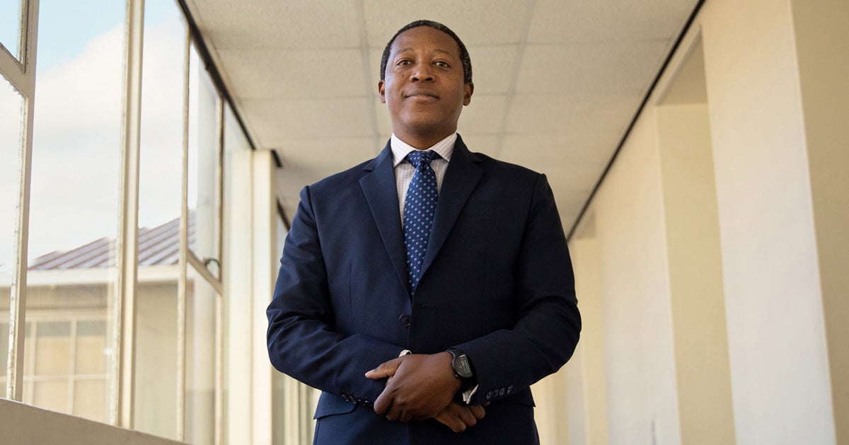 Ntobeko Ntusi stands in a hallway in a non-descript building. Windows are open behind his shoulders showing a blue sky. He wears a navy blue suit, white shirt and tie. He is black and has close-cut hair.