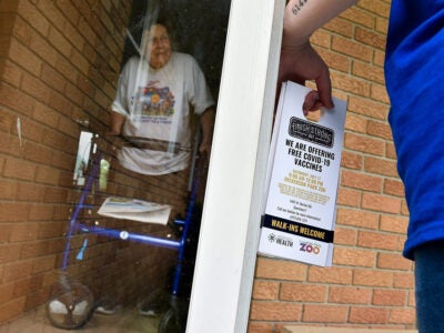 An elderly white person pushing a blue walker stands in an entryway of a yellow brick building. A white screen door is held open by a figure just barely in the frame. The person holds a door tags that say: “We are offering free covid-19 vaccines” and “walk-ins welcome.” The rest of the text is illegible.