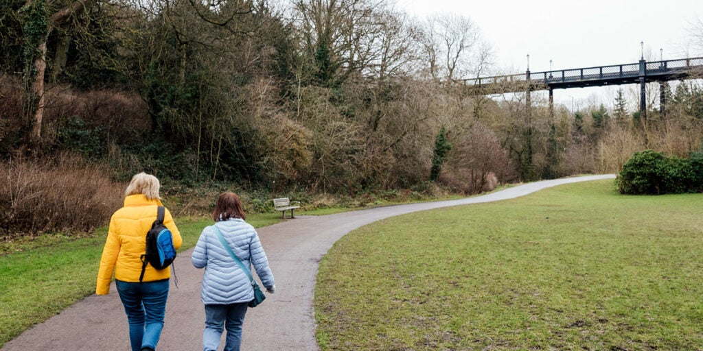 Two women wearing winter coats, jeans and bags, walk on a paved path in a nature park on a cloudy day.