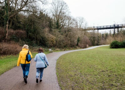Two women wearing winter coats, jeans and bags, walk on a paved path in a nature park on a cloudy day.