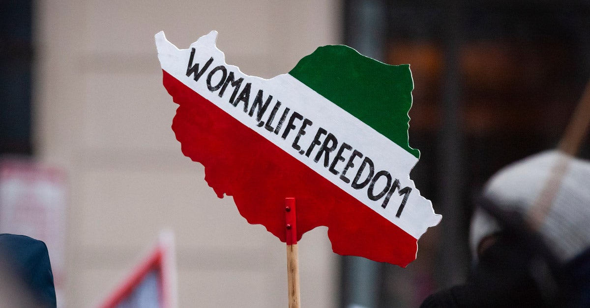 A woman holds a wooden sign of the country of Iran, painted in its flag colors: three horizontal stripes, green then white then red, at a protest. “Woman, Life, Freedom” is written in the white section.