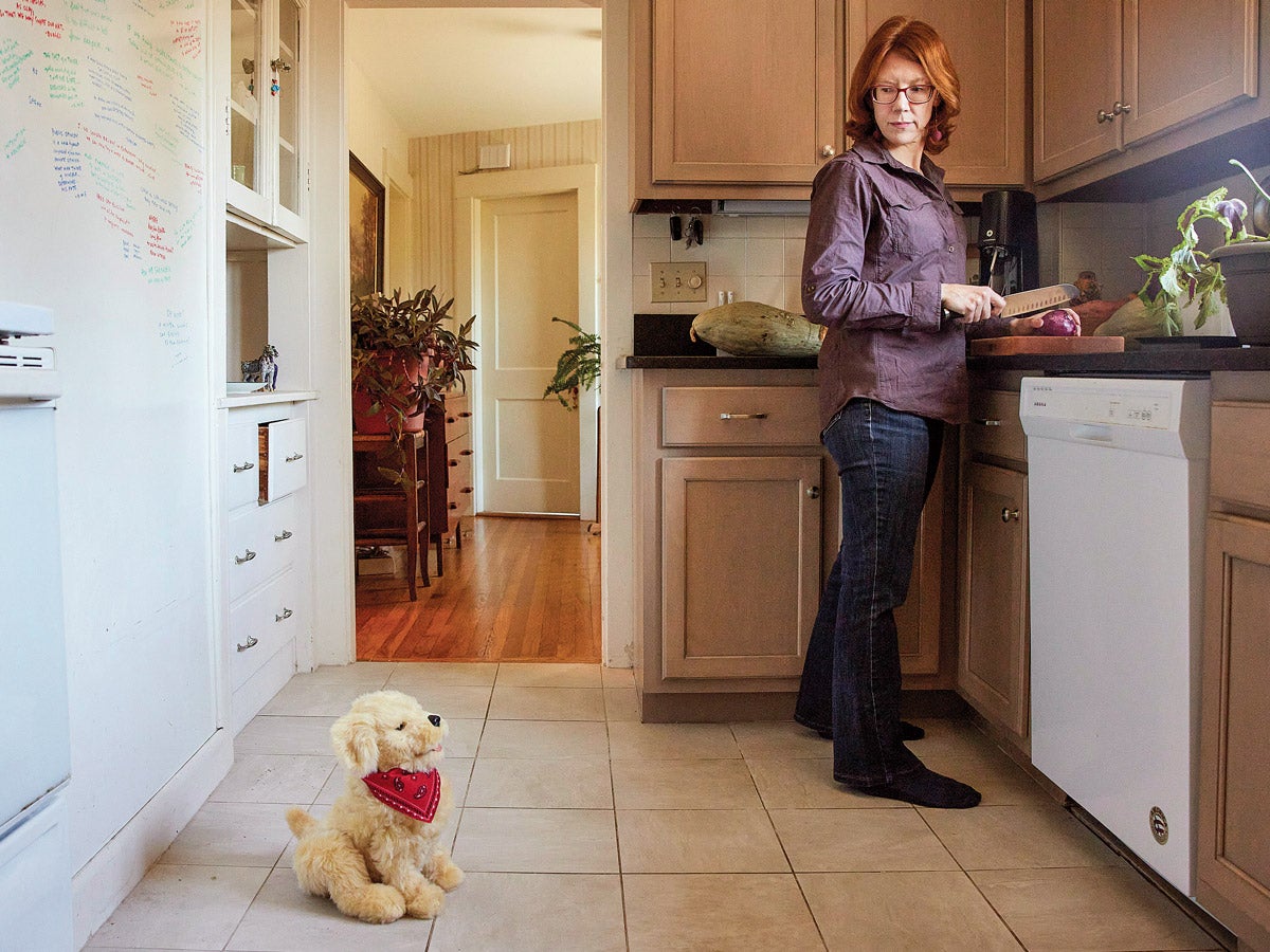 A red-headed woman wearing glasses stands in her kitchen, slicing a red onion on the counter. She peers over her shoulder at a blond, stuffed, robotic dog sitting on the white tiled floor.