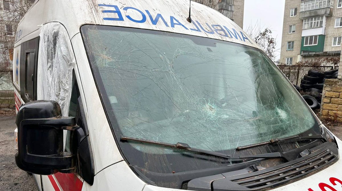 An ambulance with a windshield cracked and fractured in multiple places. It's passenger window is blown out and covered with plastic. The vehicle itself is dirty.
