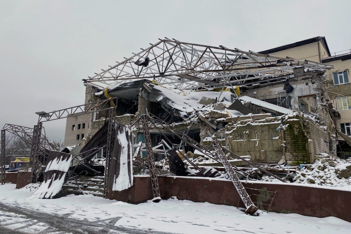 A partially destroyed hospital building is covered with snow. Exterior walls are blown out, the roof is frame-only yet parts of the concrete and yellow brick walls still stand. A short brick wall and stairs lead to the destroyed entrance.