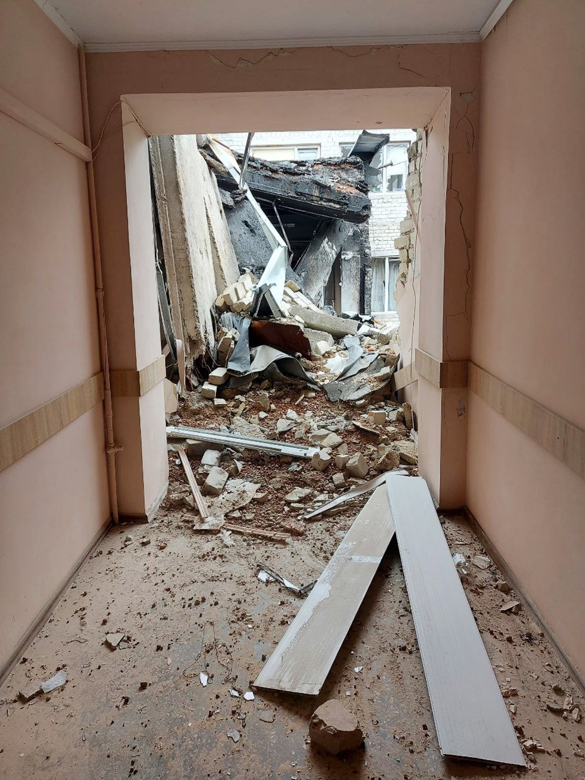 A light pink hospital hallway leads to mass destruction: fallen walls, no ceiling, debris and dirt on the floor. Two panels line flat on the floor in the hallway.