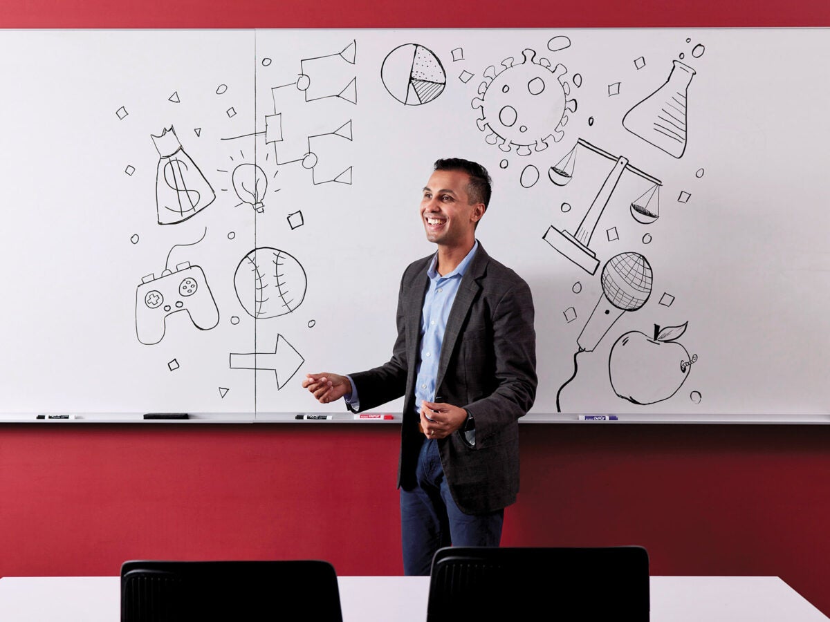 Ankur Pandya speaks in front of a whiteboard. The whiteboard has black and white illustrations of objects — a chart, scales, a COVID virus, a microphone, a baseball, arrows, a video game holder, a flow chart, beaker — that frame his figure. He looks off-camera and his hands are mid-expression.