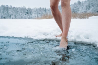 Two bare female legs step into a pond of frozen water in a snowy field.
