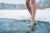 Two bare female legs step into a pond of frozen water in a snowy field.