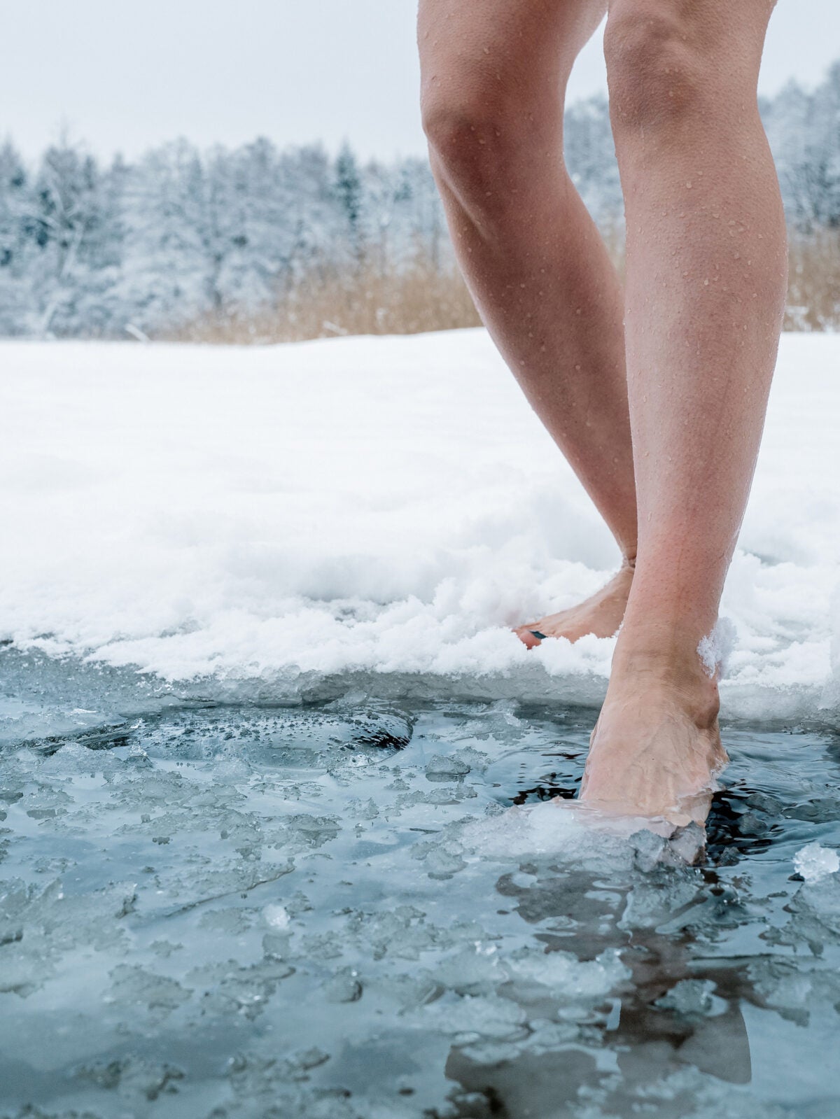 Two bare female legs step into a pond of frozen water in a snowy field.