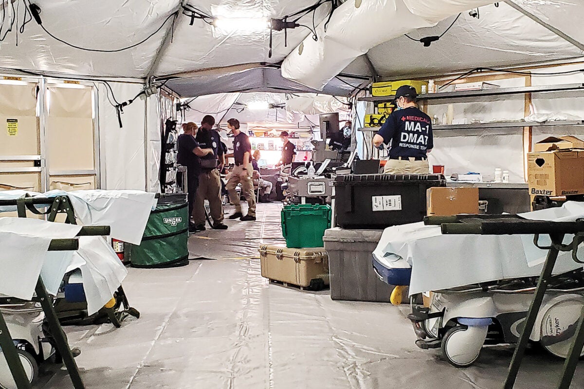 Medical workers in black t-shirts and khakis work in a large white text filled with green, black, gray, and white medical bins and portable hospital beds.