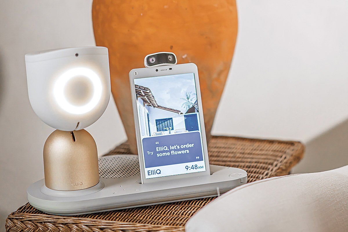 EllieQ, a desktop-sized device with a gold base and white oval shape on top, glows with a ring of white light. A tablet and camera are attached to the base on its right. EllieQ sits on a wicker side table with an orange vase in the background.