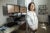 Susan Chen, wearing a lab coat, stands in her office with three computer screens displaying charts and data behind her.