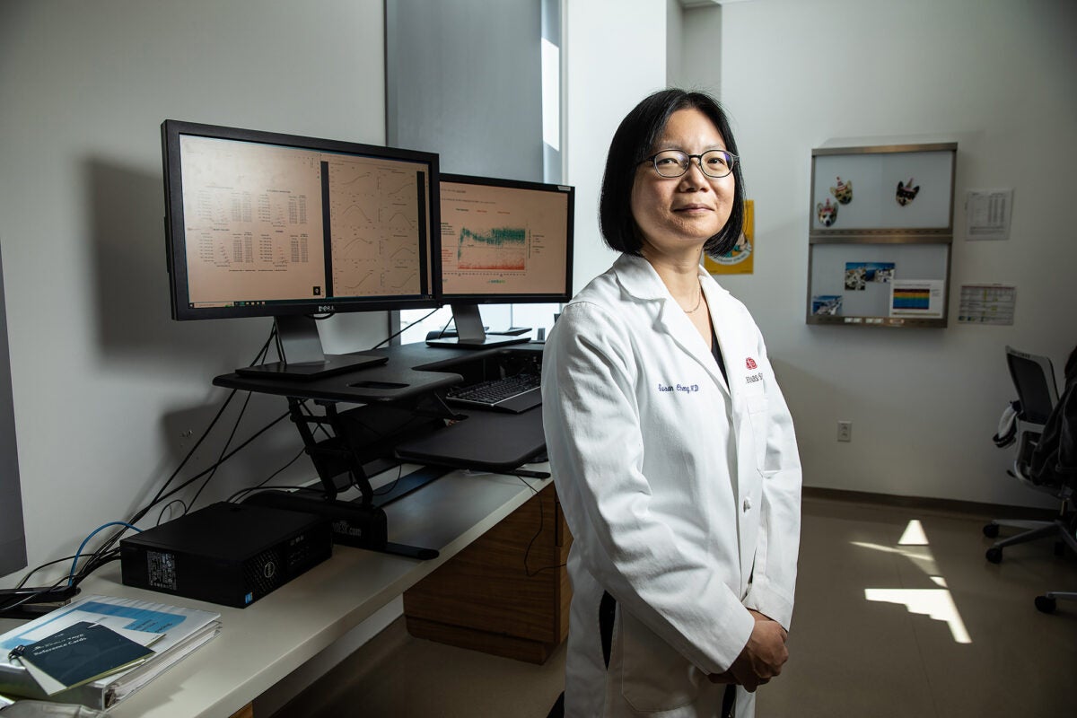 Susan Chen, wearing a lab coat, stands in her office with three computer screens displaying charts and data behind her.
