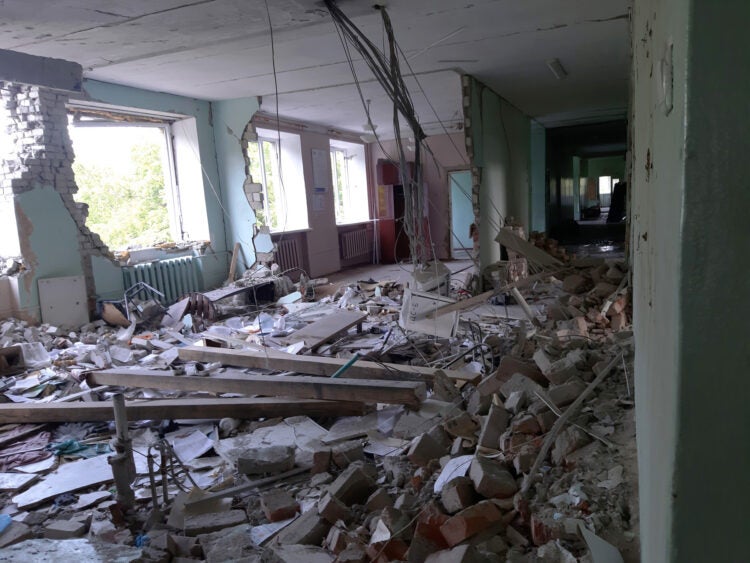 A wide shot of a third floor of a hospital building destroyed from military attacks. Debris, stone, and dirt are everywhere. Windows are blown out, wires hang from the ceiling.