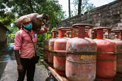 West Bengal, India: A man wears a pink button-down shirt, green cloth mask, and head wrap while holding a large canister of cooking gas next to a truck bed with multiple canisters.