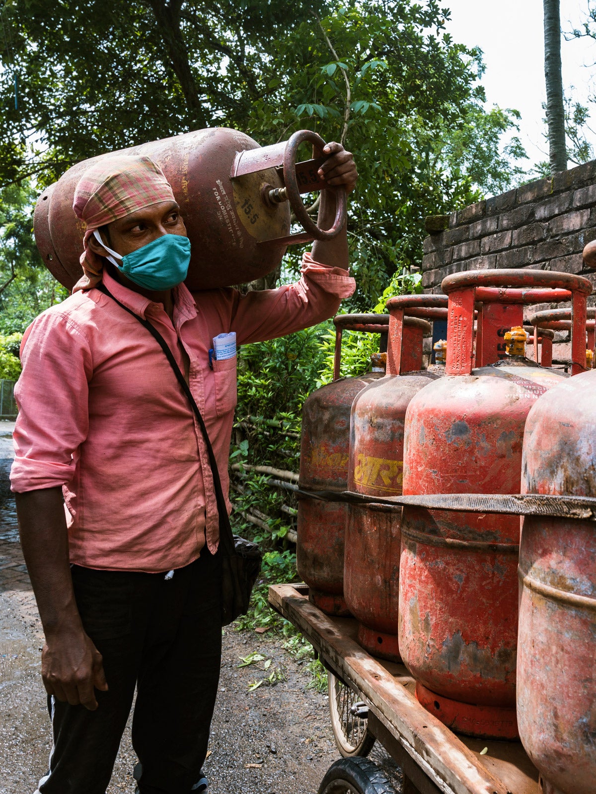 West Bengal, India: A man wears a pink button-down shirt, green cloth mask, and head wrap while holding a large canister of cooking gas next to a truck bed with multiple canisters.