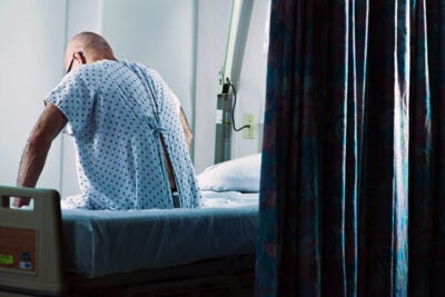 A bald, adult man wearing a hospital gown sits on the side of a hospital bed with his back facing the camera. His hands rest beside him in a pensive, thinking, position. A hospital curtain is in the foreground.