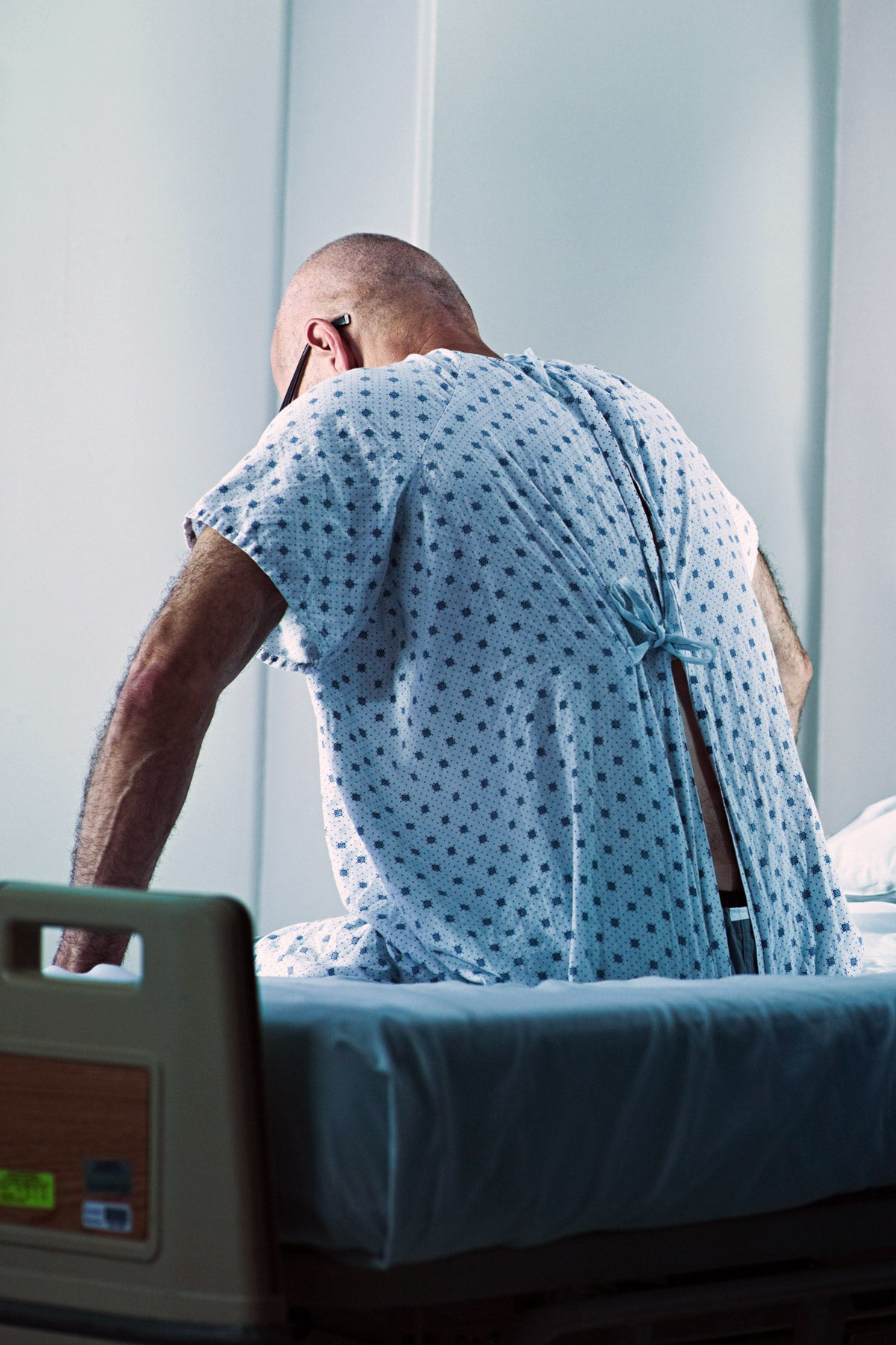 A bald, adult man wearing a hospital gown sits on the side of a hospital bed with his back facing the camera. His hands rest beside him in a pensive, thinking, position. A hospital curtain is in the foreground.