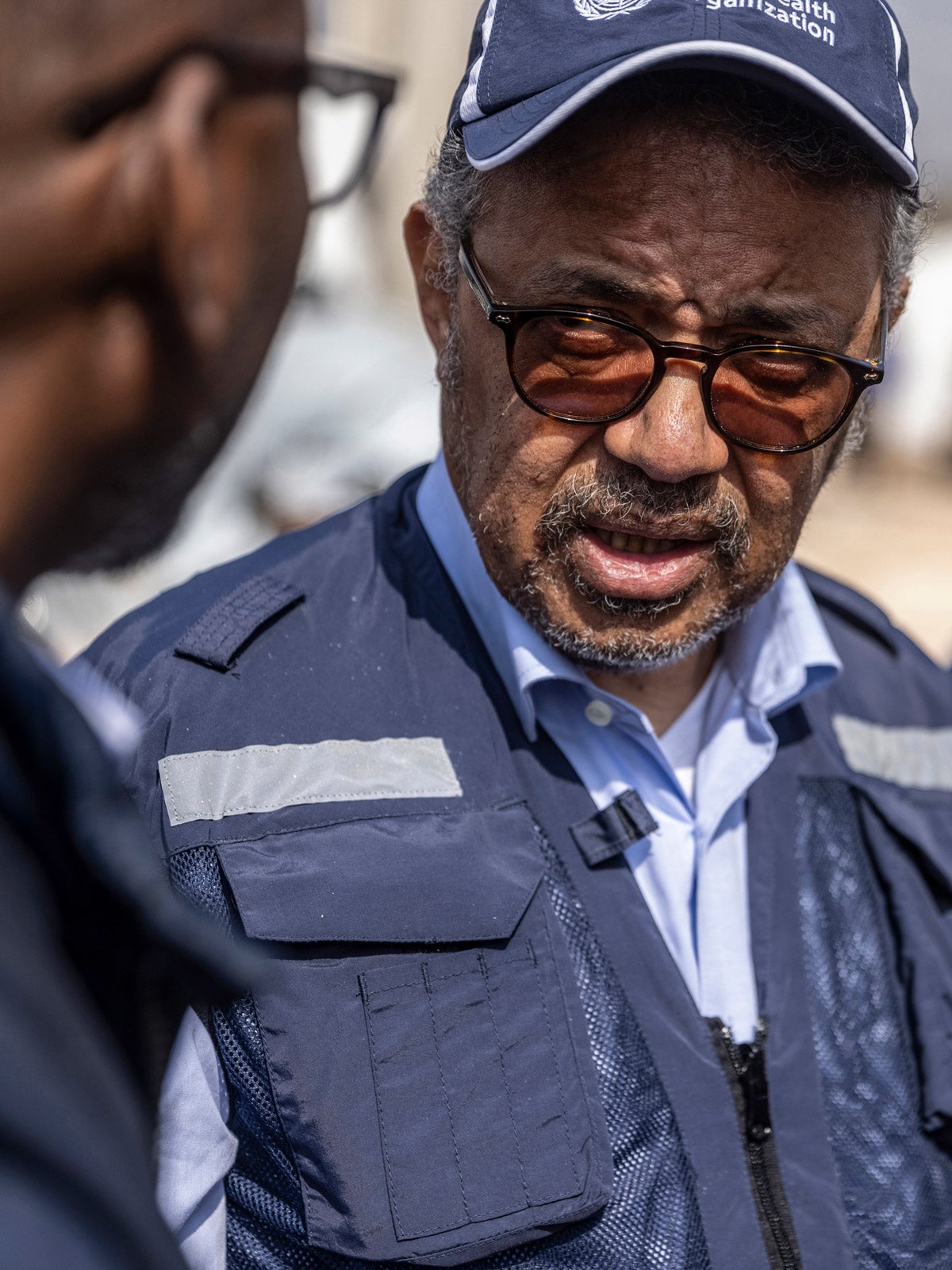 Director-General of the World Health Organization, Tedros Adhanom Ghebreyesus, wearing a dark blue World Health Organization (WHO) aid vest and hat speaks to a figure off-camera. He is standing outside at a border crossing near the Syran-Turkish border.
