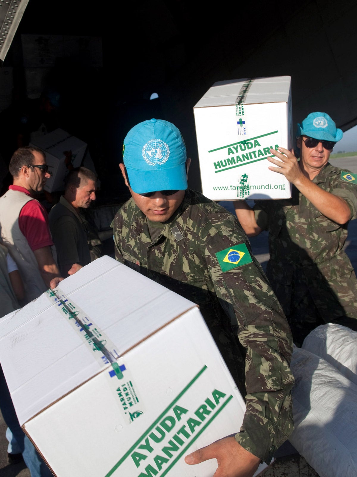 An aid officer, wearing camouflage with a Brazilian flag path and a blue WHO baseball cap, carries a white box with the words “Ayuda Humanitarian” written in green. Another man carries one behind him. They are on a tarmac and are carrying the boxes around a landed plane. Other aid workers are in the background.