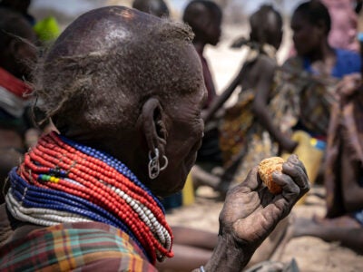 A woman of the East African Turkana tribe eats the seed of a palm tree. She is mostly bald, wears traditional tribal clothing and a beaded necklace and a silver earring made of a can tab. The palm seed is brown-red, is round, shriveled and about the size of a clementine.
