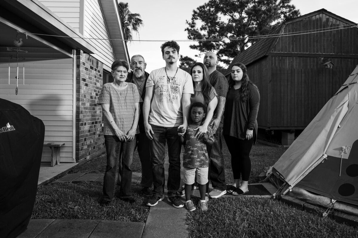 Black and white photo: Seven family members stand close together and stare directly into the camera. In the center is a twenty-something man, who hols the hand of a young black boy. To his left are an elderly man and woman. To his right are a middle-aged woman who holds the young boys other hand, a middle-aged man, and a young woman. They stand in a backyard, with a grill and house on one side, and a modern tipi on the other.