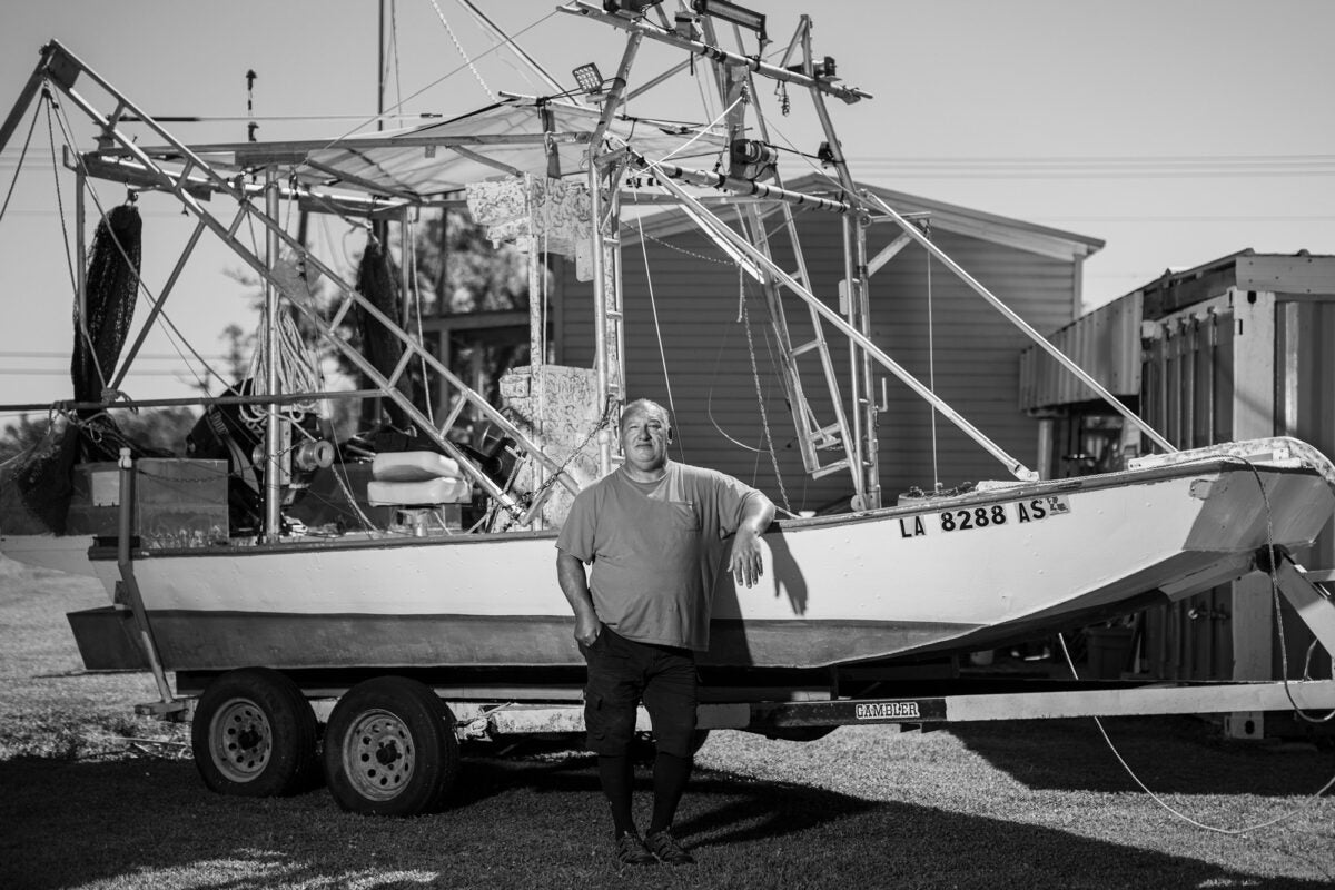 Black and white photo: A bald, middle aged man stands in front a fishing boat on a trailer, resting his arm on the side. He stares directly at the camera.
