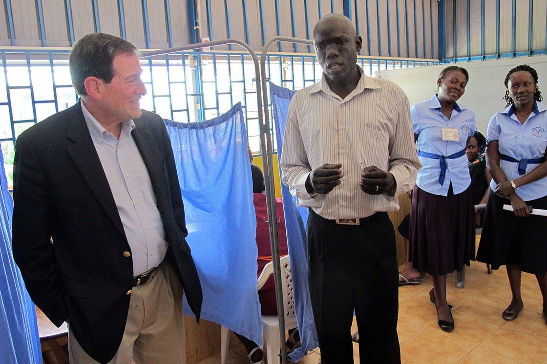 Archival photo, 2012: A tall African man wearing a dress shirt and pants speaks to a white man wearing a dress shirt, pants and blazer in an African clinic. Two clinic workers stand behind them.