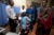Hillary Clinton stands in an Ugandan clinic, and is greeted by a family. A woman kneels in front of her and holds a toddler, a male stands with a child, and three clinic officials stand beside her.