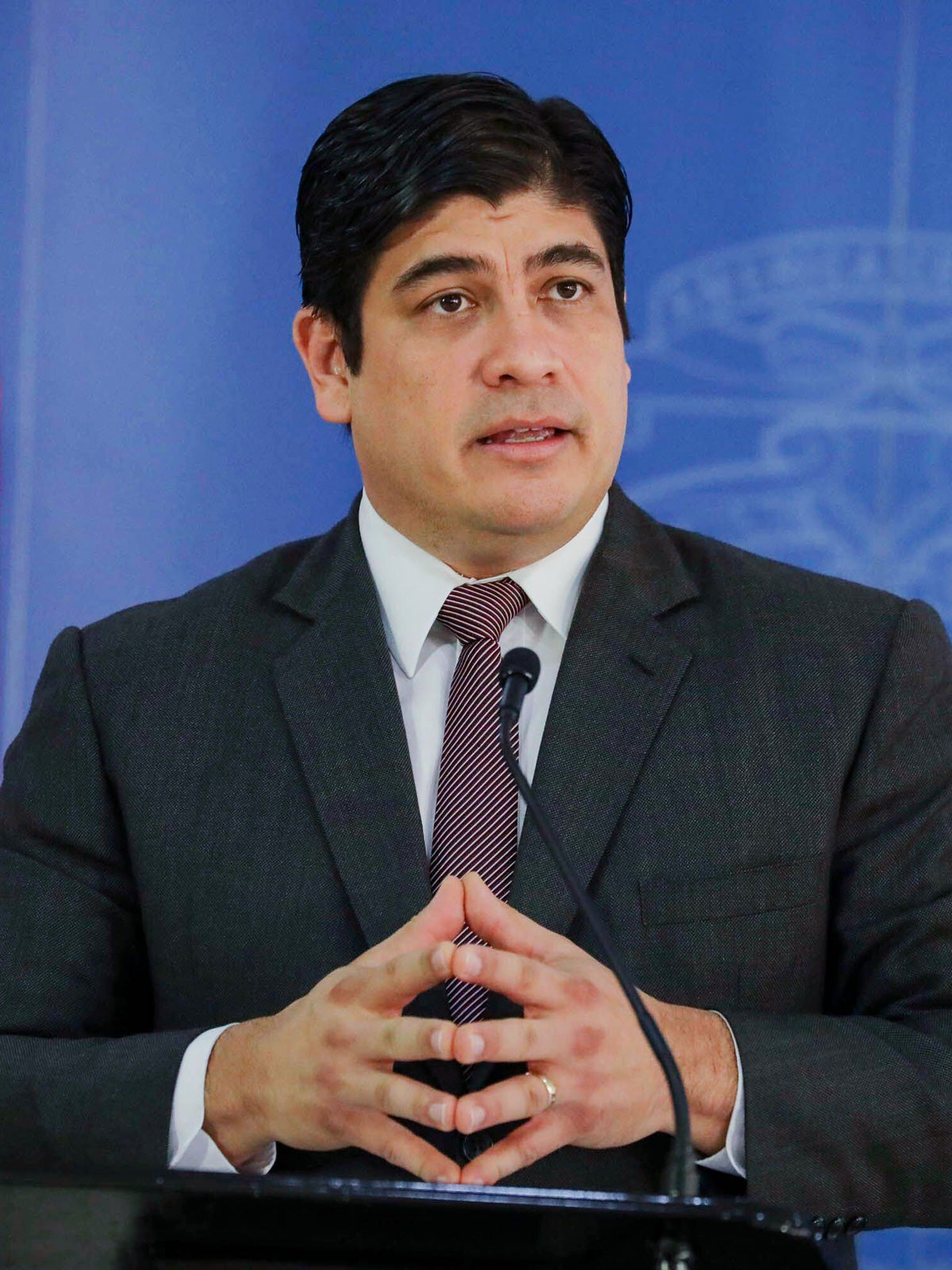 Carlos-Alvarado-Quesada speaks at a podium with his hands tented. He wears a dark suit and tie and is in front of a blue background.