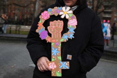 A female wearing a black winter jacket holds a cardboard sign in the shape of the female symbol that is decorated with colorful, hand-drawn flowers and a fist raised in the middle.