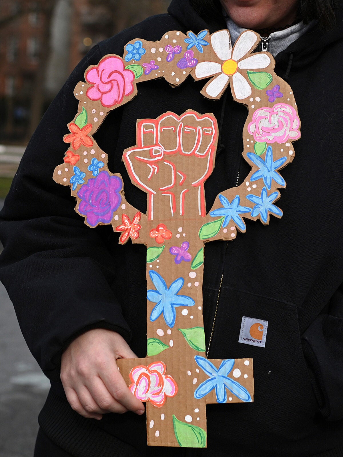 A female wearing a black winter jacket holds a cardboard sign in the shape of the female symbol that is decorated with colorful, hand-drawn flowers and a fist raised in the middle.