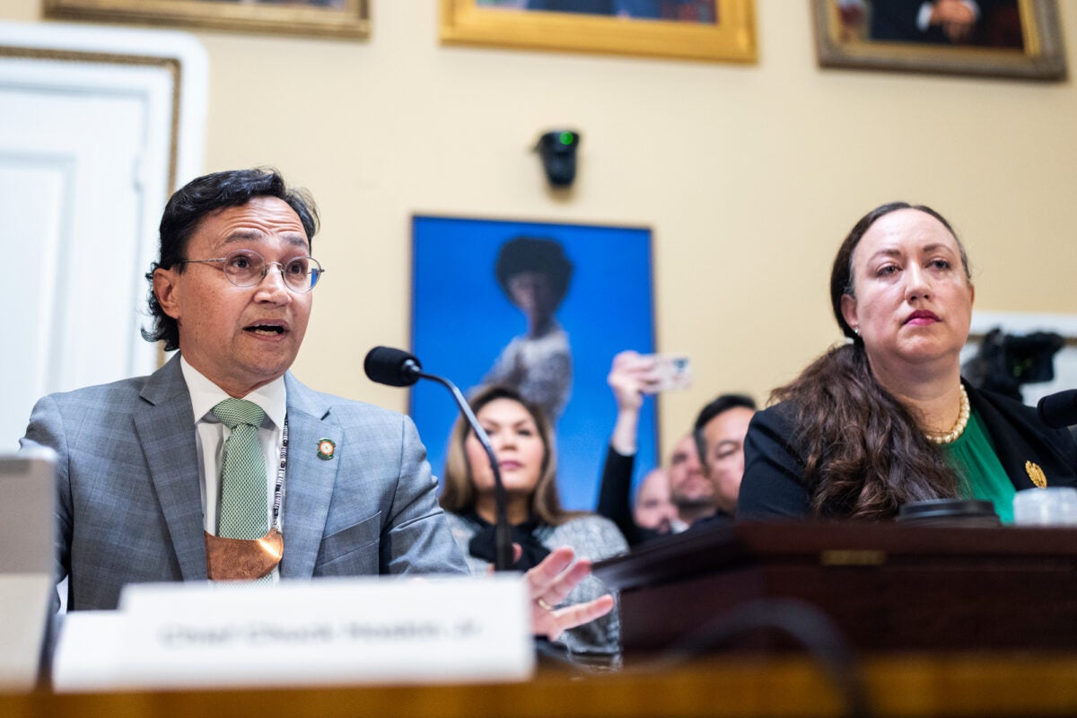 Chief Chuck Hoskin Jr., chief of the Cherokee Nation, and Mainon Schwartz, attorney at the Congressional Research Service, sit behind a table with microphones and namecards at a House Rules Committee. Chief Hoskin wears a light suit, tie, and a traditional necklace with a large bronze pendant. Schwartz wears a blue blazer, gold pendant, teal shirt, and peals.