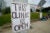 A poster board with the words “This clinic is open” in all-caps hangs from a pole in front of a green lawn and pink building, a women’s health clinic.