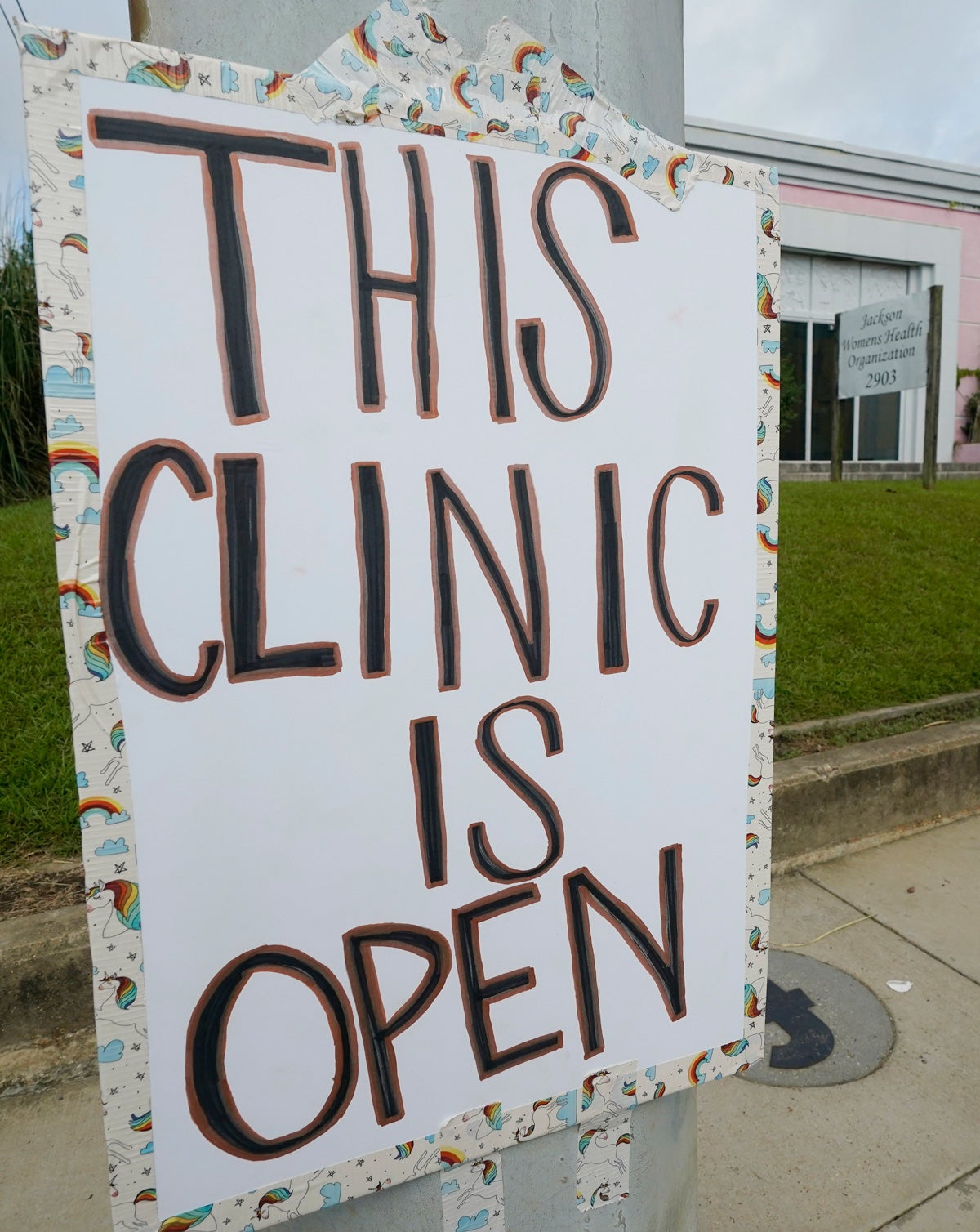 A poster board with the words “This clinic is open” in all-caps hangs from a pole in front of a green lawn and pink building, a women’s health clinic.