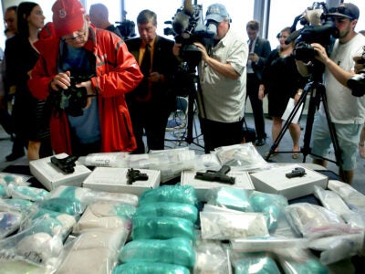 A group of photographers, videographers, and members of the press stand over a table taking photos of seized handguns, fentanyl, heroin, and cocaine from a bust of a major drug trafficking ring.